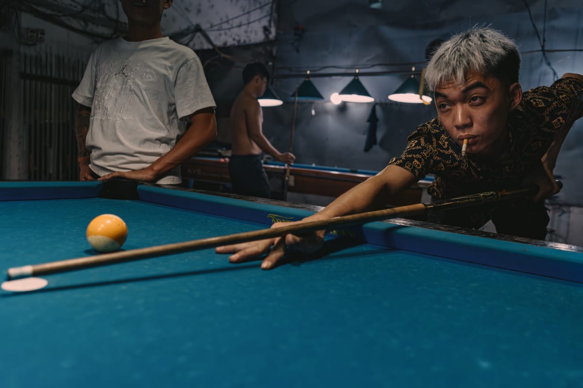 A grey-haired man with a cigarette in his lips stretches across a blue table to strike a yellow ball in a dim pool hall — Antonio di Vico