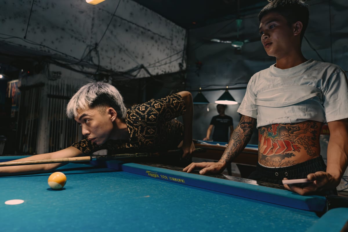 A silver-haired player sights low along his cue at a yellow ball while a tattooed man in a cropped white shirt looks on — Antonio di Vico