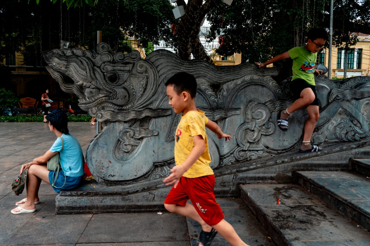A boy in a yellow shirt and red shorts runs past a carved stone dragon balustrade as another child in green climbs it and a woman rests nearby — Antonio di Vico