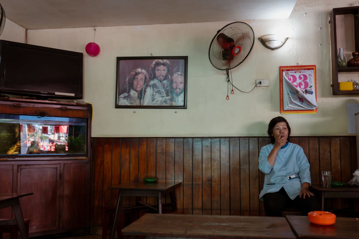 A woman in a pale blue jacket smokes at a wooden-paneled cafe beneath a framed Bee Gees portrait, a wall fan, and a glowing fish tank — Antonio di Vico