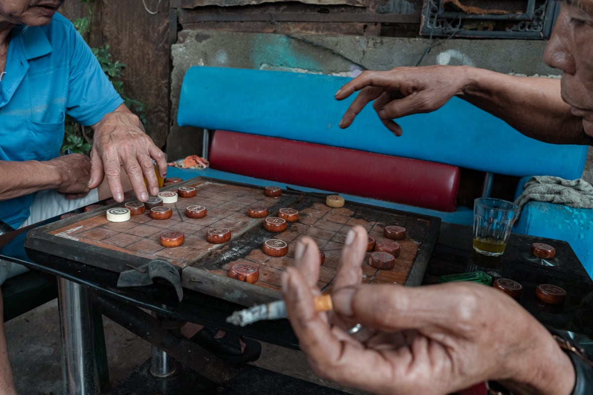 Weathered hands of two elderly men reach over a worn wooden Chinese chess board, one holding a lit cigarette, beside a glass of tea outdoors — Antonio di Vico