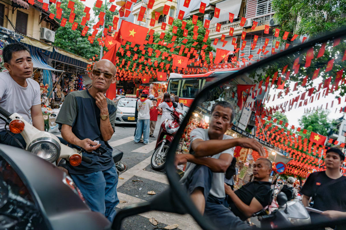 A bespectacled man pauses beneath dense red Vietnamese flag bunting in a busy market street, his companion reflected in a scooter's side mirror — Antonio di Vico