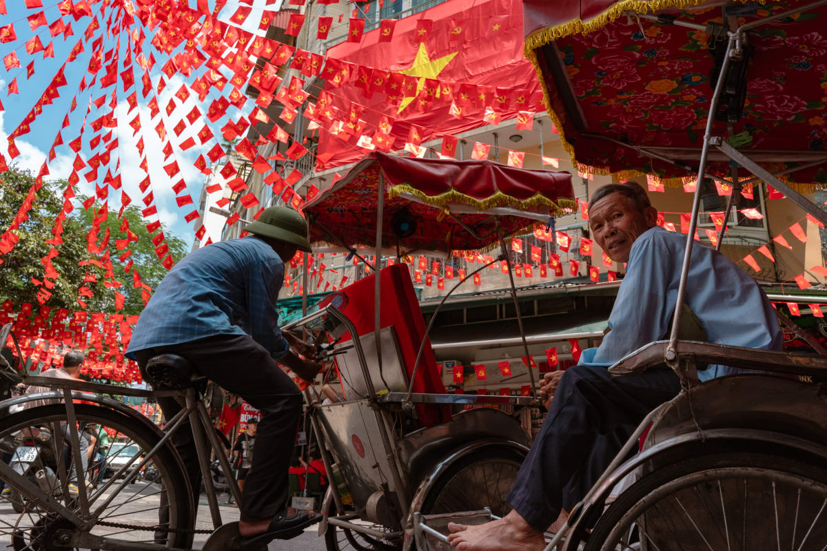 Two older men rest on a cyclo beneath a dazzling canopy of red Vietnamese and Communist Party flags against blue sky — Antonio di Vico