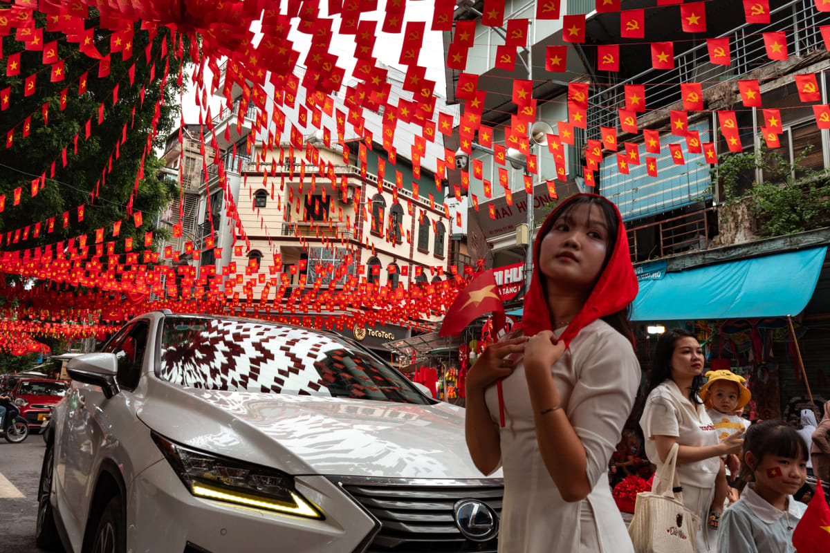 A young woman in a white ao dai holds a small national flag beneath a thick red flag canopy as a silver Lexus and families pass on the festive street — Antonio di Vico