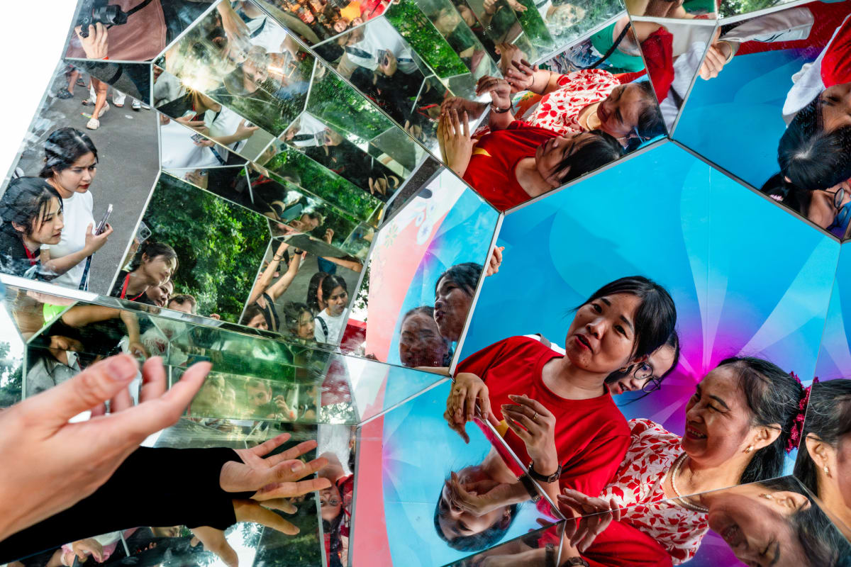 Vietnamese women in red crowd a mirrored kaleidoscope sculpture, their faces and reaching hands fractured into bright geometric reflections — Antonio di Vico