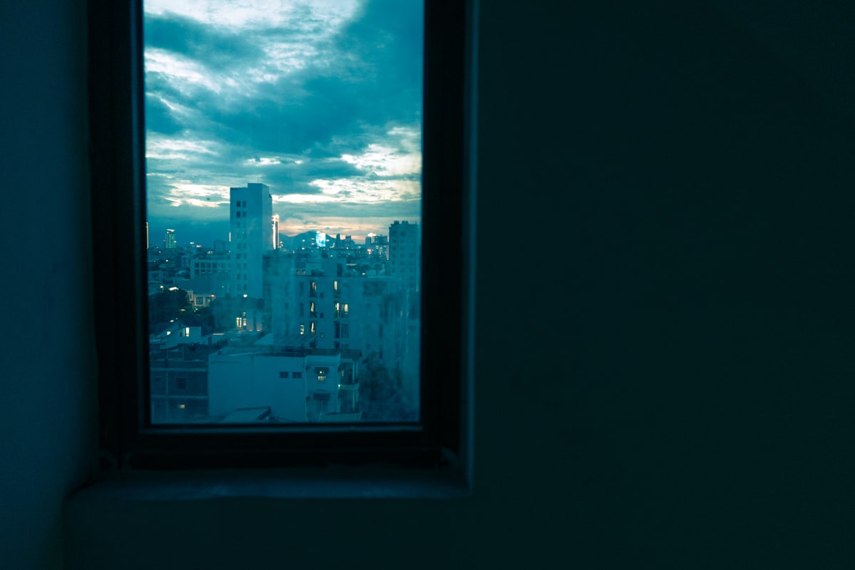 Through a dark room's window, a teal dusk sky glows over Hanoi's low rooftops and lit apartment towers — Antonio di Vico