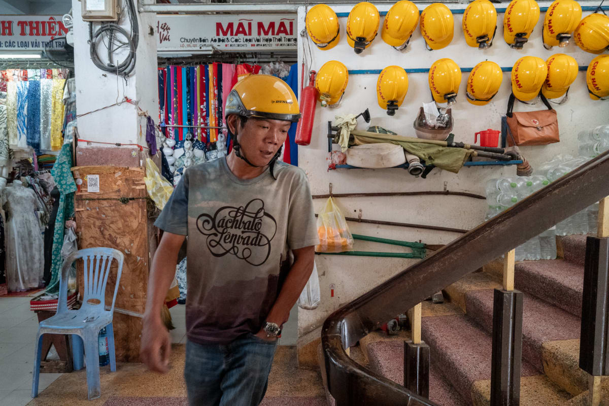 A man in a yellow Dunlop motorbike helmet walks past a fabric shop and a wall of yellow safety helmets beside a stairwell — Antonio di Vico