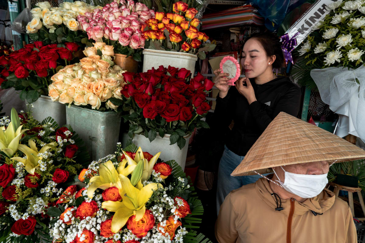 A woman checks a pink hand mirror amid buckets of red, pink, and orange roses and lilies as a masked vendor in a conical hat passes — Antonio di Vico