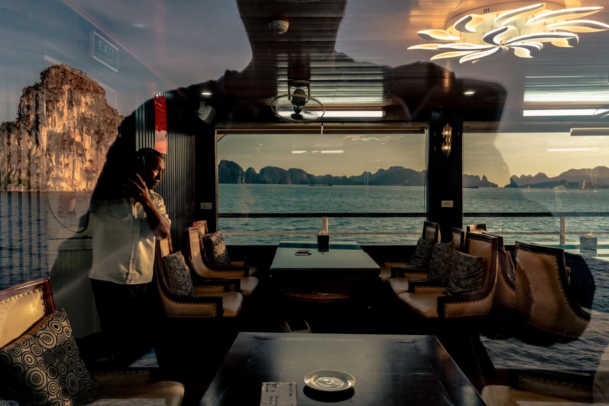 Reflections layer a cruise boat's empty leather dining lounge over Ha Long Bay's karst islands, a headphoned crew member at left — Antonio di Vico
