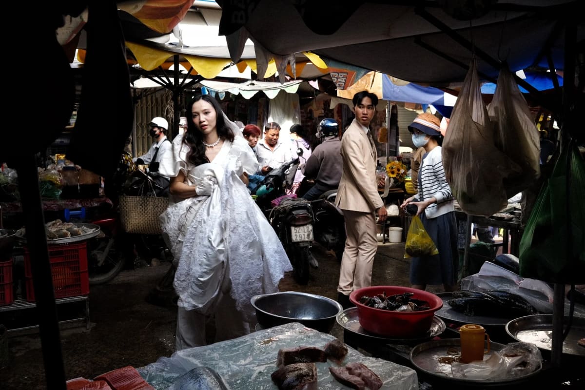 A bride gathers her white gown and a groom in a pale suit stand among the fish basins and hanging plastic bags of a crowded wet market — Luke Neville