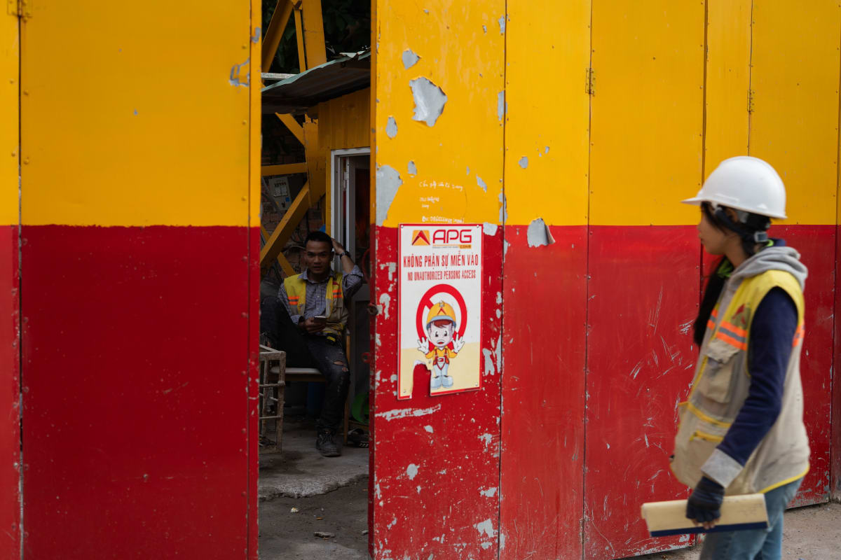 A worker in a hard hat passes a yellow-and-red construction gate as a crew rests in the doorway behind — Luke Neville