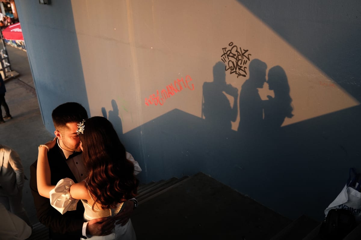 A kissing couple in wedding dress cast doubled shadows across a sunlit graffiti wall — Luke Neville