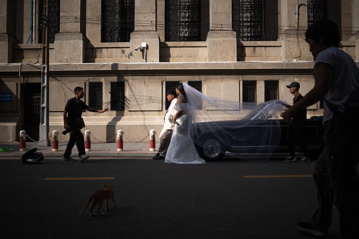 A bride's veil streams over a vintage car as attendants stretch it across the street below a stone facade — Luke Neville