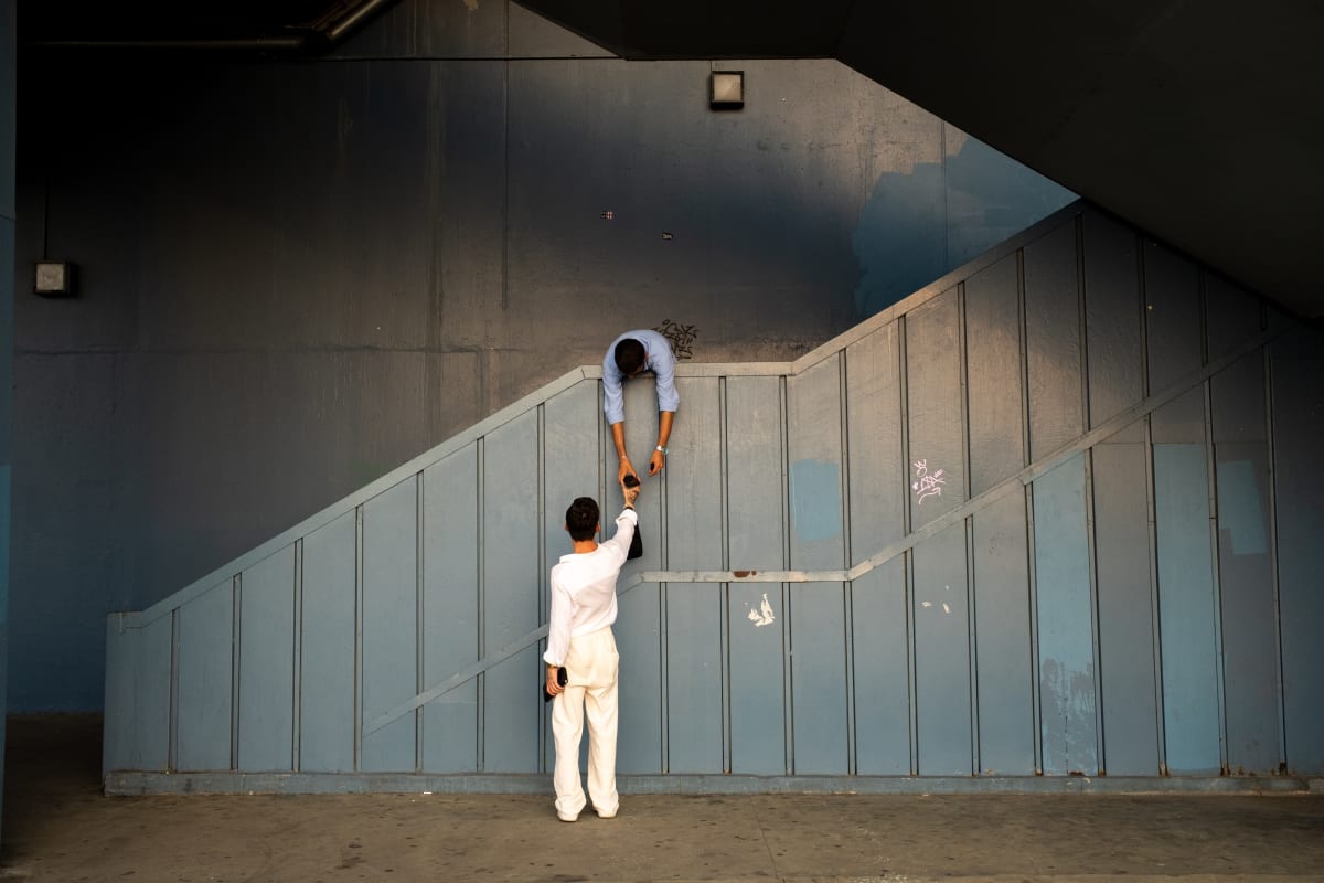 In a blue underpass, a man leans over the stair rail to pass something down to a woman in white — Luke Neville