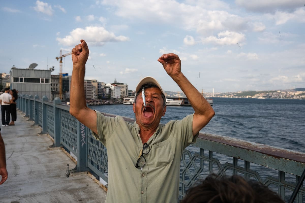 A man throws his arms up mid-shout on Istanbul's Galata Bridge, the water stretching behind him — Luke Neville