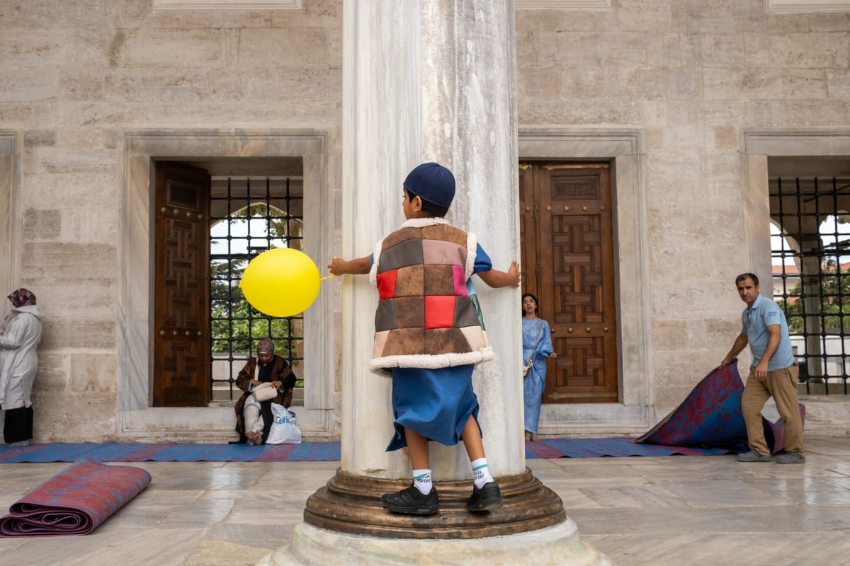 A boy holds a yellow balloon beside a marble column in a mosque courtyard as prayer rugs are rolled away behind him — Luke Neville