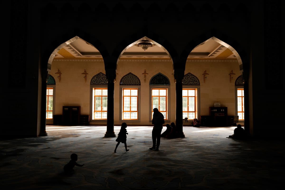 Children and worshippers move in silhouette beneath the arches of a glowing mosque hall — Luke Neville