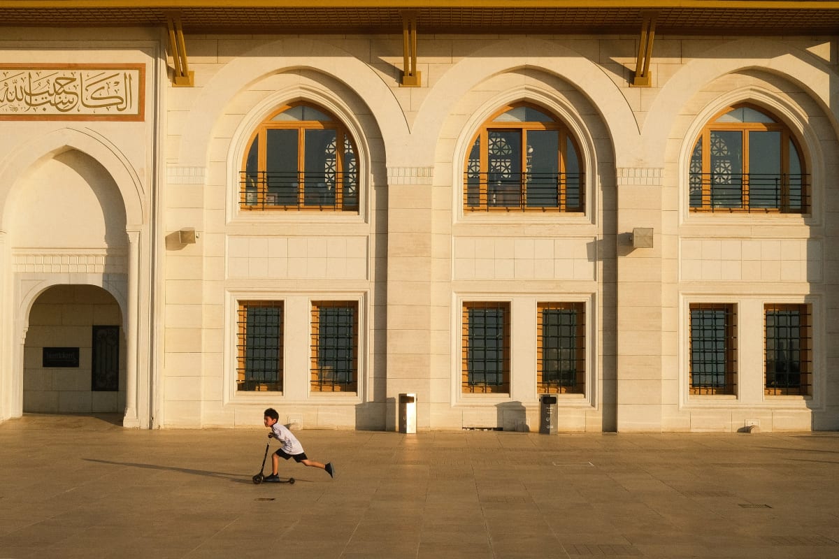 A small boy kicks his scooter across an empty mosque courtyard beneath tall arched windows — Luke Neville