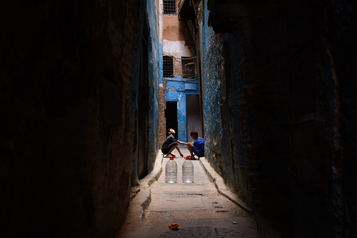 Two boys crouch with plastic water jugs at the bright end of a narrow blue alley — Luke Neville