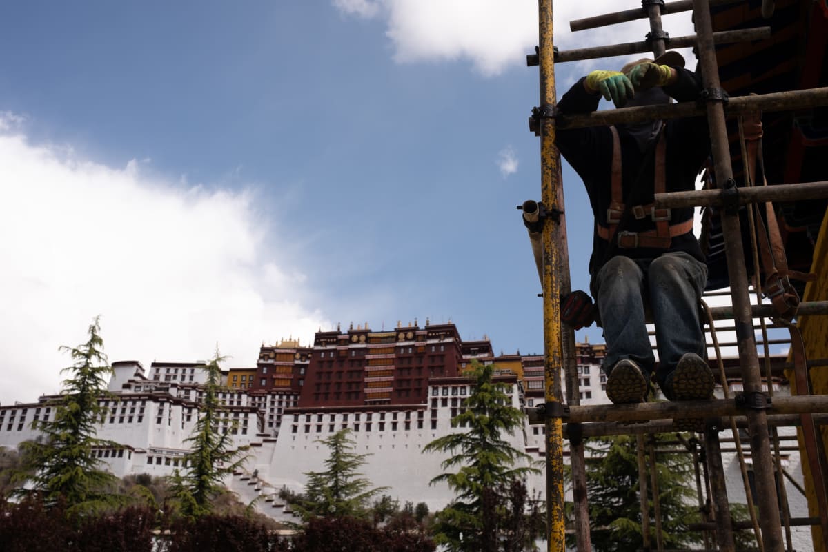A worker climbs scaffolding, the Potala Palace rising on the hill behind him — Luke Neville