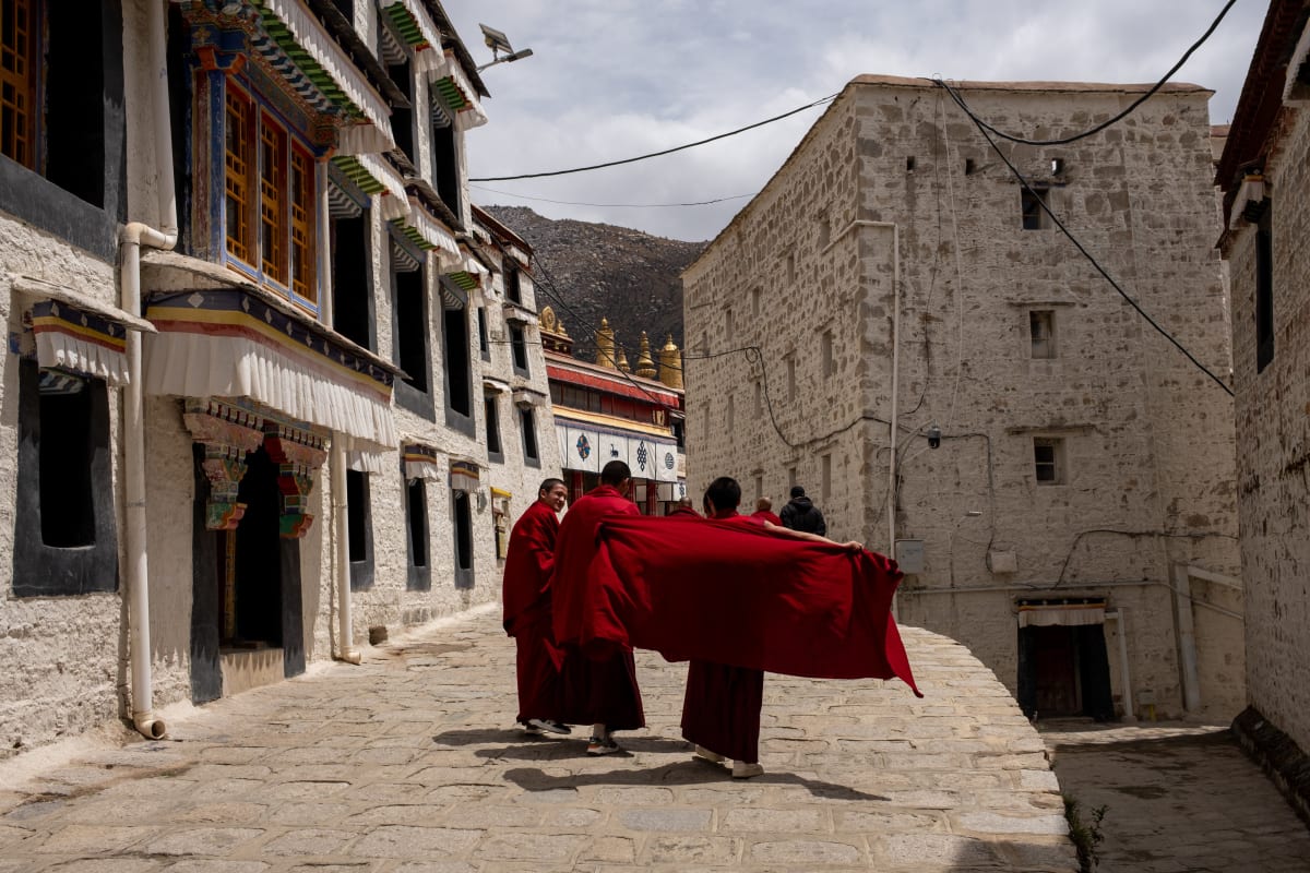 Two monks carry a billowing length of red cloth down a stone lane between whitewashed monastery walls — Luke Neville