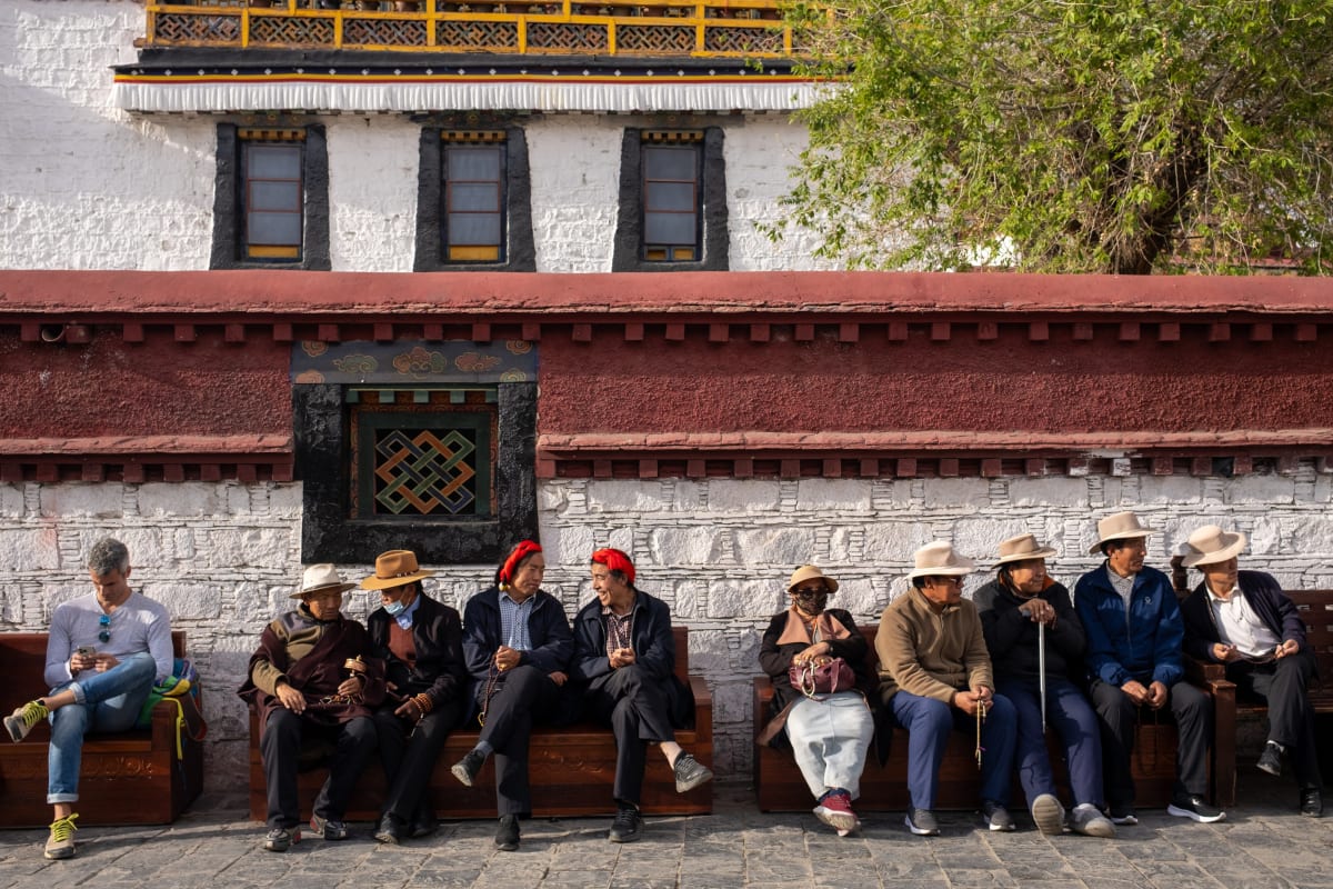 A row of elders in hats share a bench and conversation along a monastery wall — Luke Neville