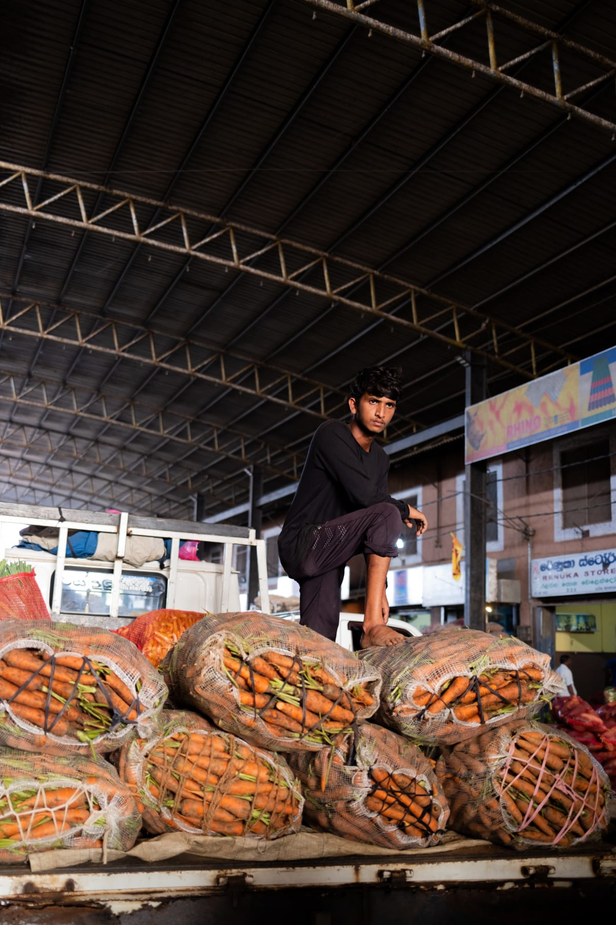 A young porter stands over netted sacks of carrots in a covered wholesale market, torch in hand — Luke Neville