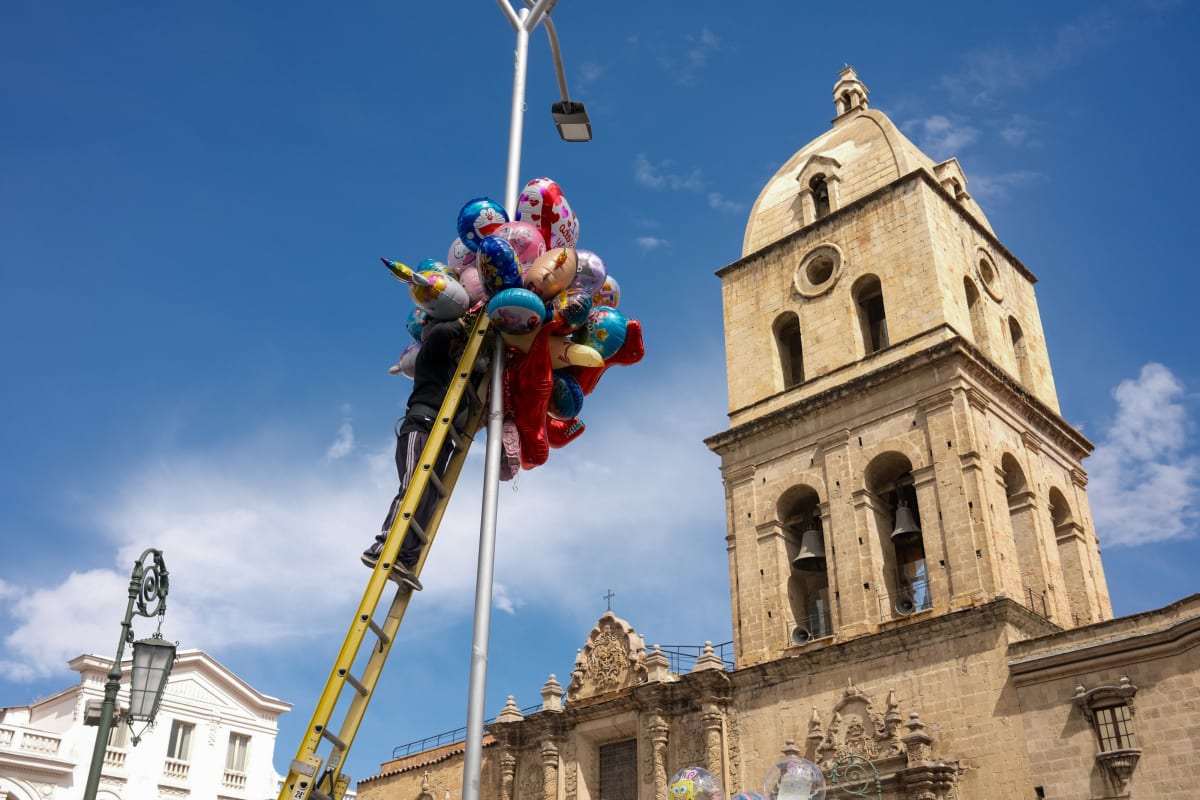 A balloon seller climbs a ladder up a lamppost, his bundle of balloons floating beside a stone bell tower in La Paz — Luke Neville