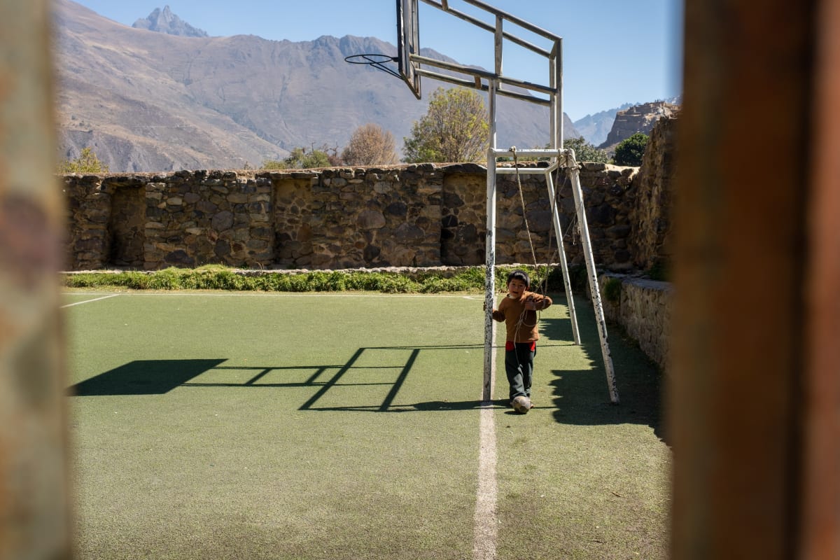 A child swings on the frame of a basketball hoop on a walled court, Andean peaks rising beyond — Luke Neville