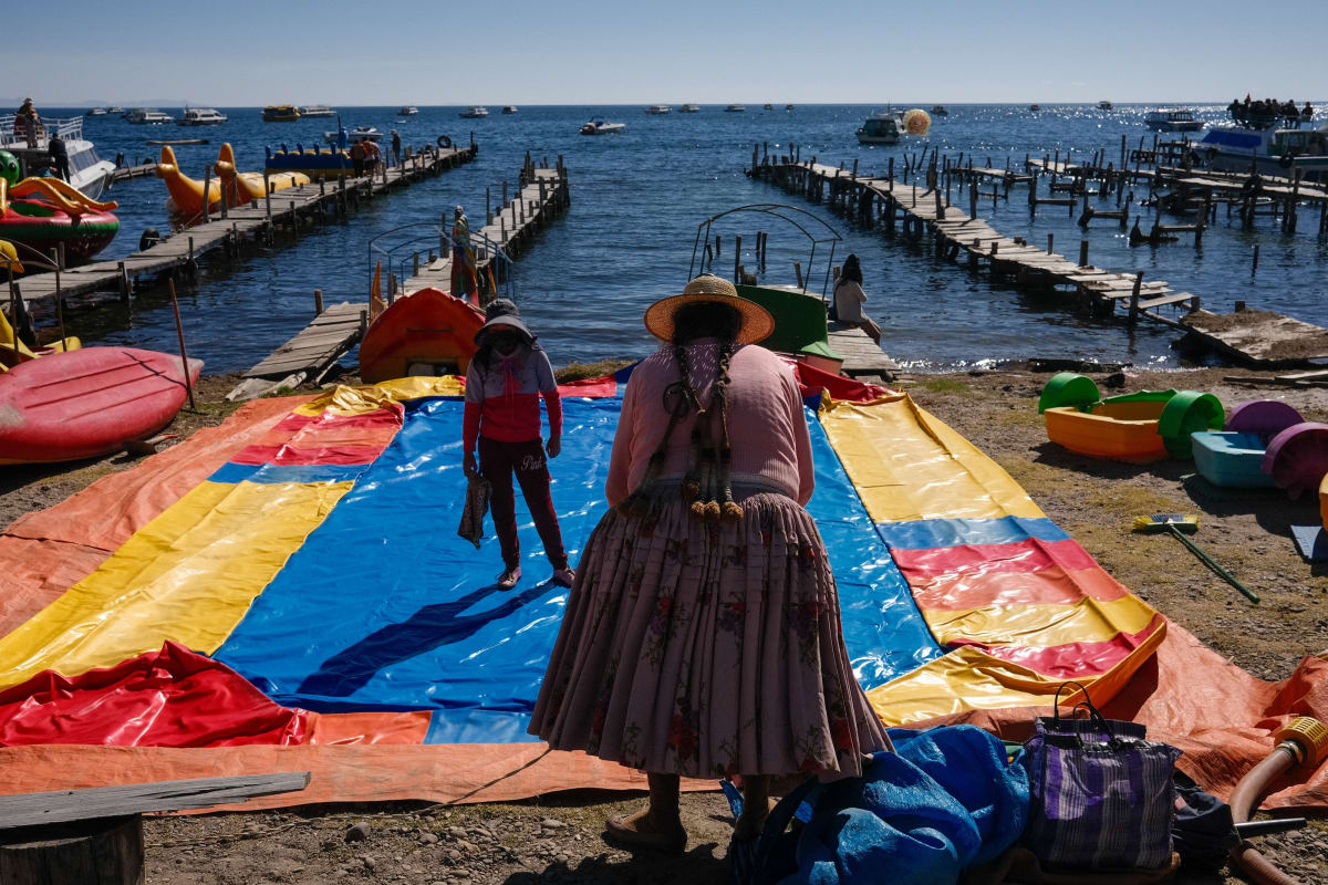 A woman in a pollera skirt stands over an inflatable slide among the pedal boats and piers of Lake Titicaca — Luke Neville