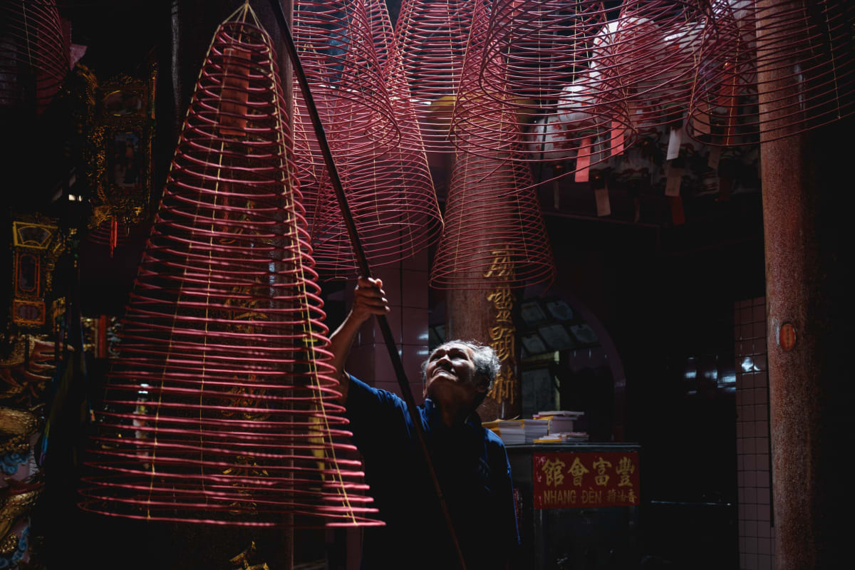 A man reaches up with a long pole to tend towering red spiral incense coils hanging in a dim Cho Lon temple — Adrien Jean