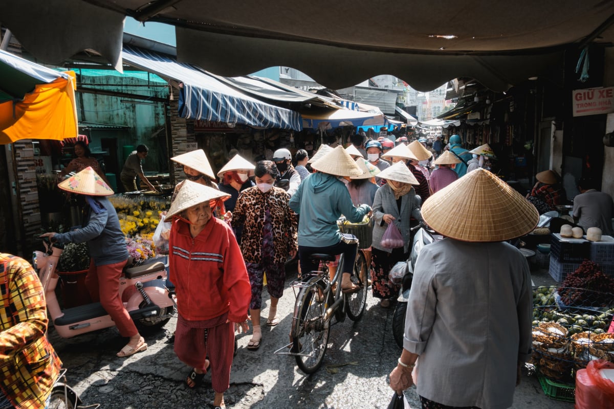 Shoppers in conical hats and a cyclist thread through a crowded, sunlit morning market lane — Adrien Jean