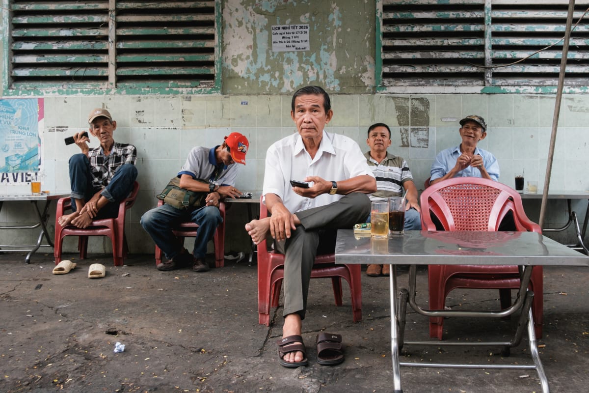 Five older men sit on red plastic chairs at an open-air café against a weathered louvred wall, one meeting the camera — Adrien Jean
