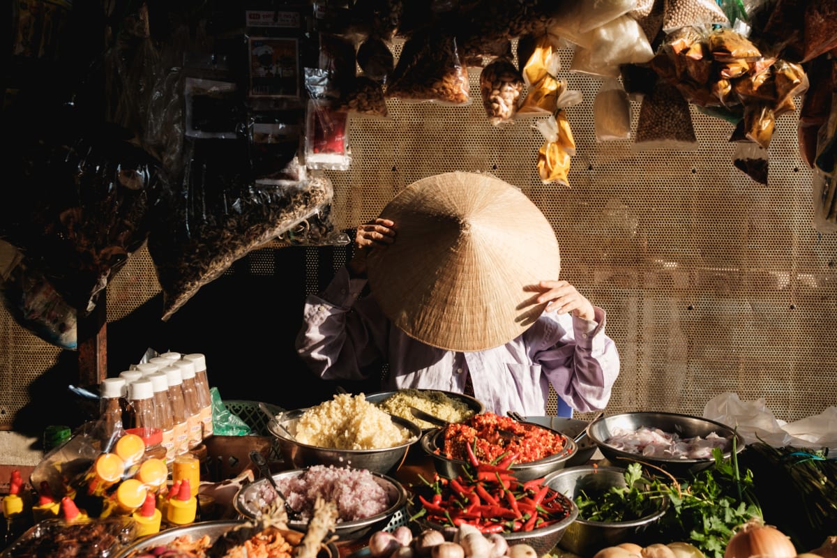 A vendor under a conical hat sits behind bowls of chilies, herbs and rice at a market food stall — Adrien Jean