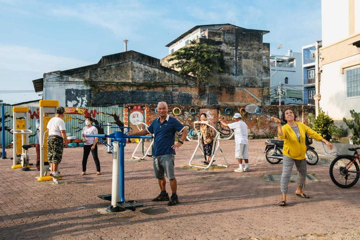 Residents use public outdoor exercise machines in a paved yard beside a graffiti-marked wall — Adrien Jean
