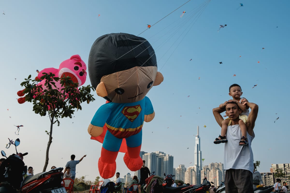 A giant inflatable Superman kite floats above a riverside kite field as a man carries a child on his shoulders, Landmark 81 beyond — Adrien Jean