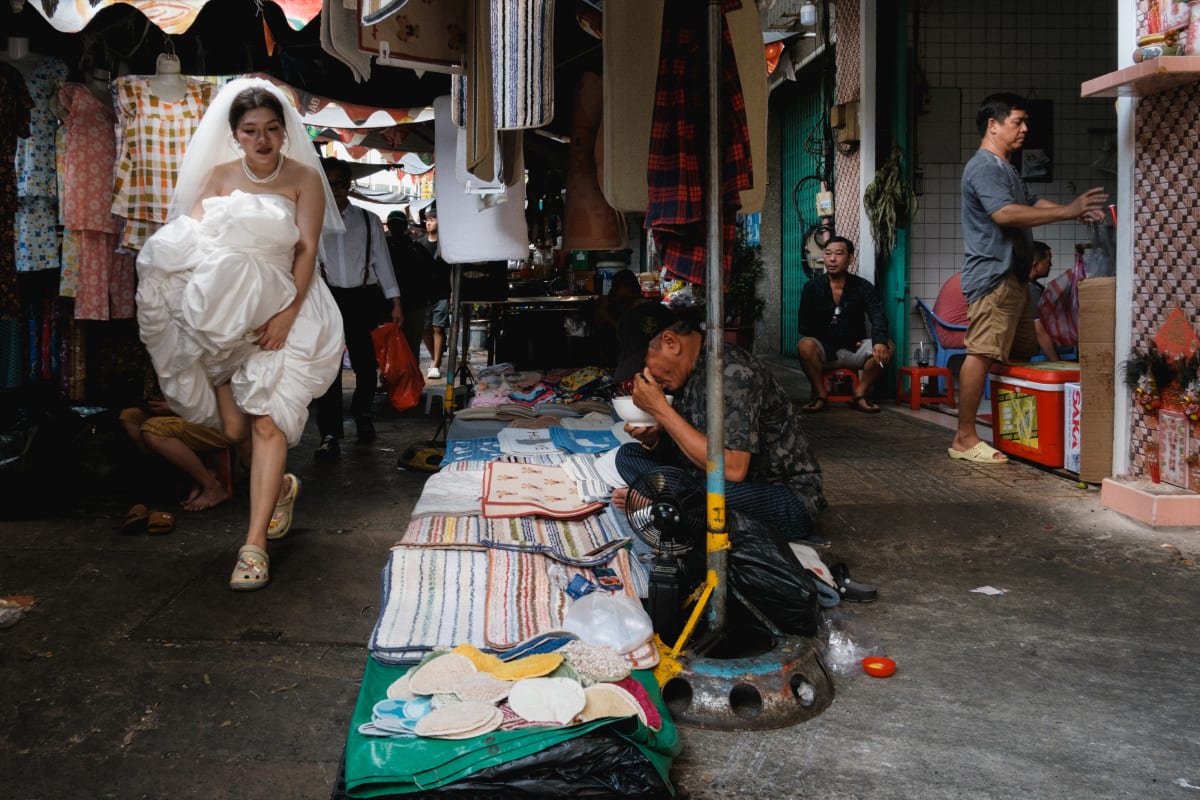 A bride in a white gown walks through a market lane, passing a vendor seated on the ground — Adrien Jean