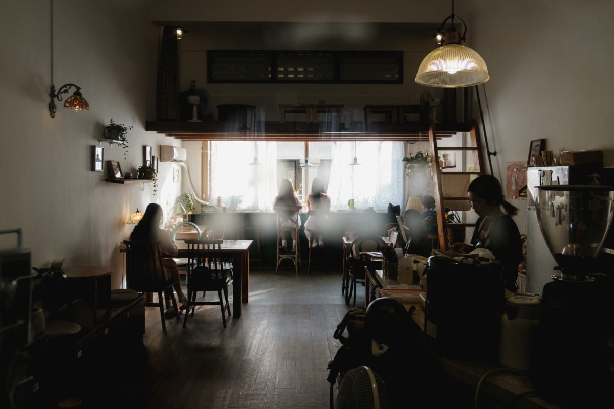 Patrons sit in a quiet café interior, soft daylight falling through backlit windows — Adrien Jean