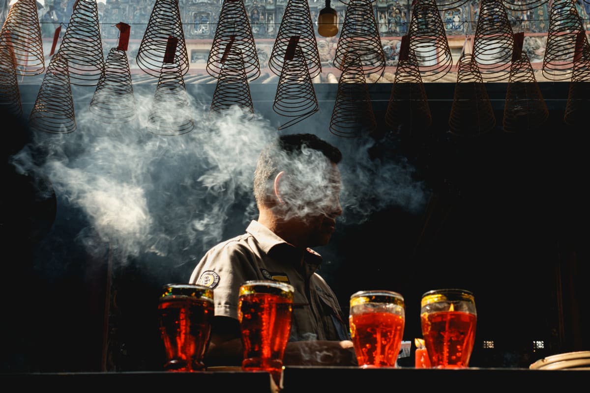 A man's silhouette rises through incense smoke behind a row of backlit tea glasses, spiral coils overhead — Adrien Jean