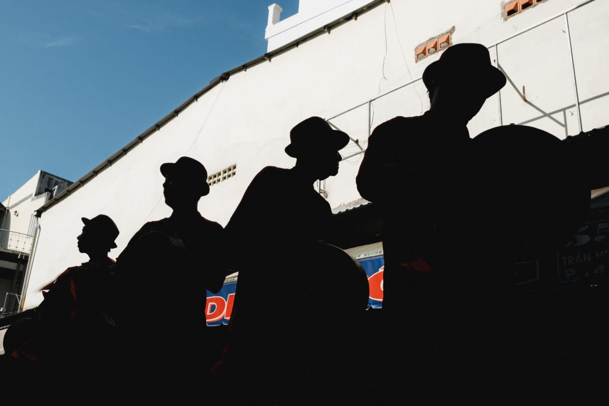 The hard-edged silhouettes of four men in hats line up against a bright white wall — Adrien Jean