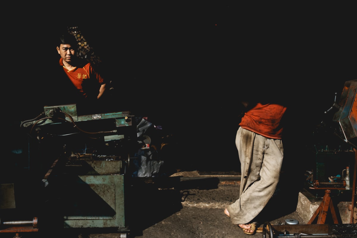 In a dim metal workshop, a boy works at a machine while another figure in red crosses the floor — Adrien Jean