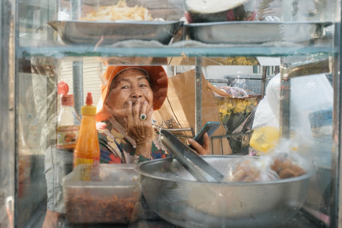 Seen through the glass of her food cart, a street vendor rests her chin on her hand and smiles — Adrien Jean