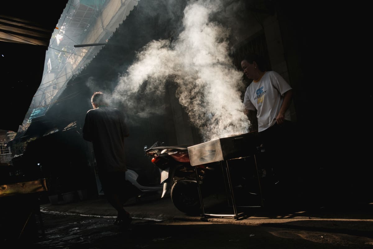 Steam billows through a shaft of light at a night street-food stall as a vendor works beside a parked motorbike — Adrien Jean