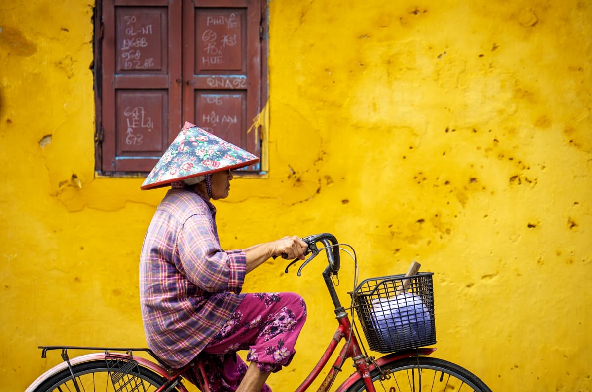 A woman in a floral conical hat cycles a red bicycle past a weathered yellow wall and a shuttered maroon window in Hoi An — Jim Kerns