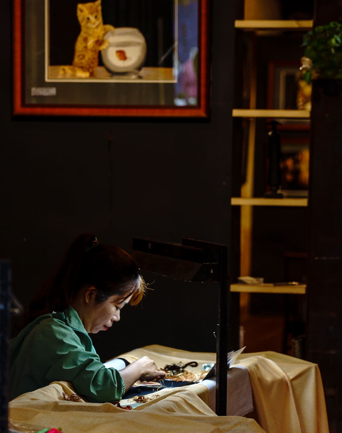 A woman in a green shirt bends over her embroidery frame in a dark workshop, a framed picture of a kitten on the wall above her — Jim Kerns