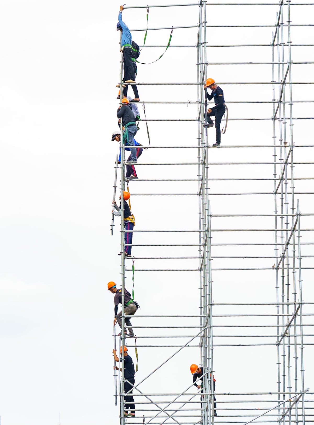Workers in harnesses and orange helmets climb a lattice of steel scaffolding against a blank white sky — Jim Kerns