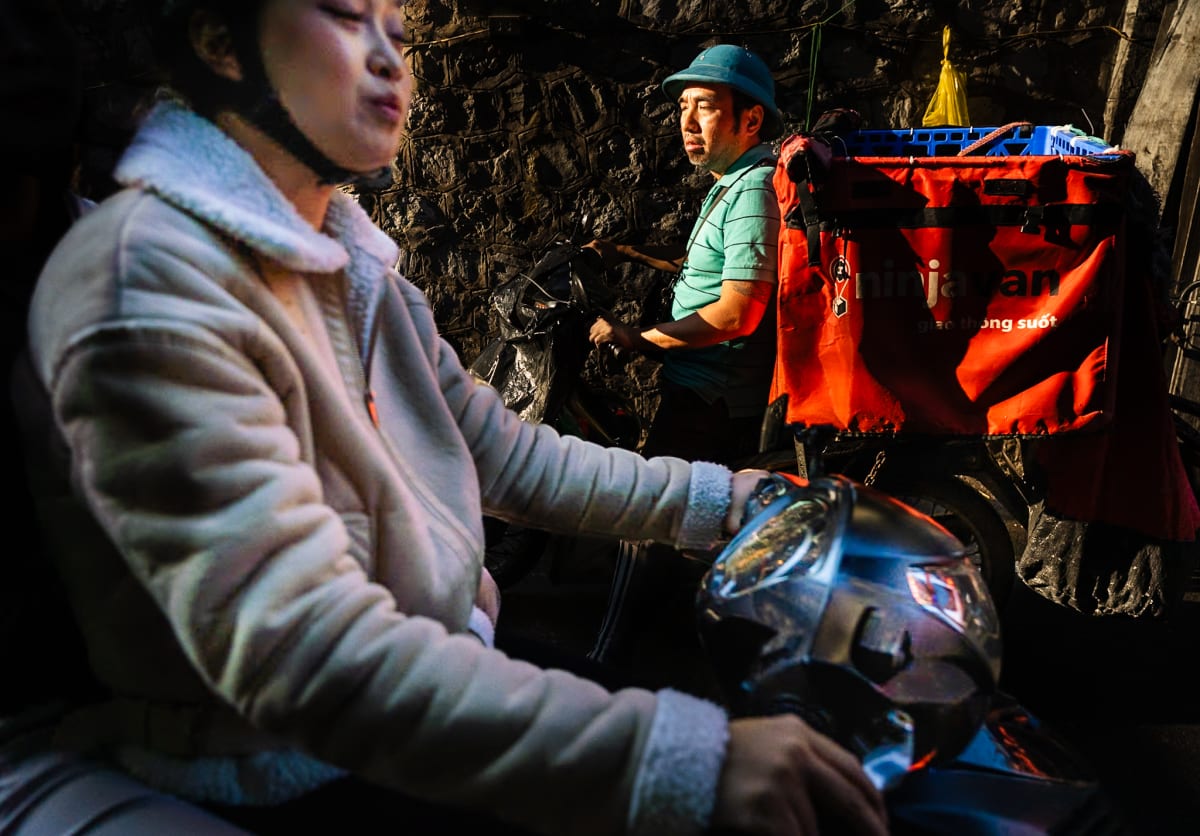 A woman on a scooter and a delivery rider with an orange cargo box pass one another in a dark stone alley — Jim Kerns