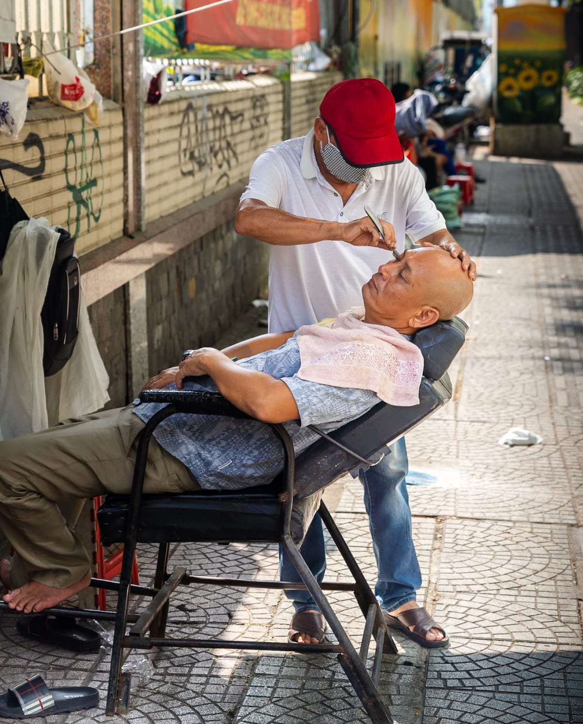 A barber in a red cap shaves a reclining customer's scalp on a folding chair set up on the pavement — Jim Kerns