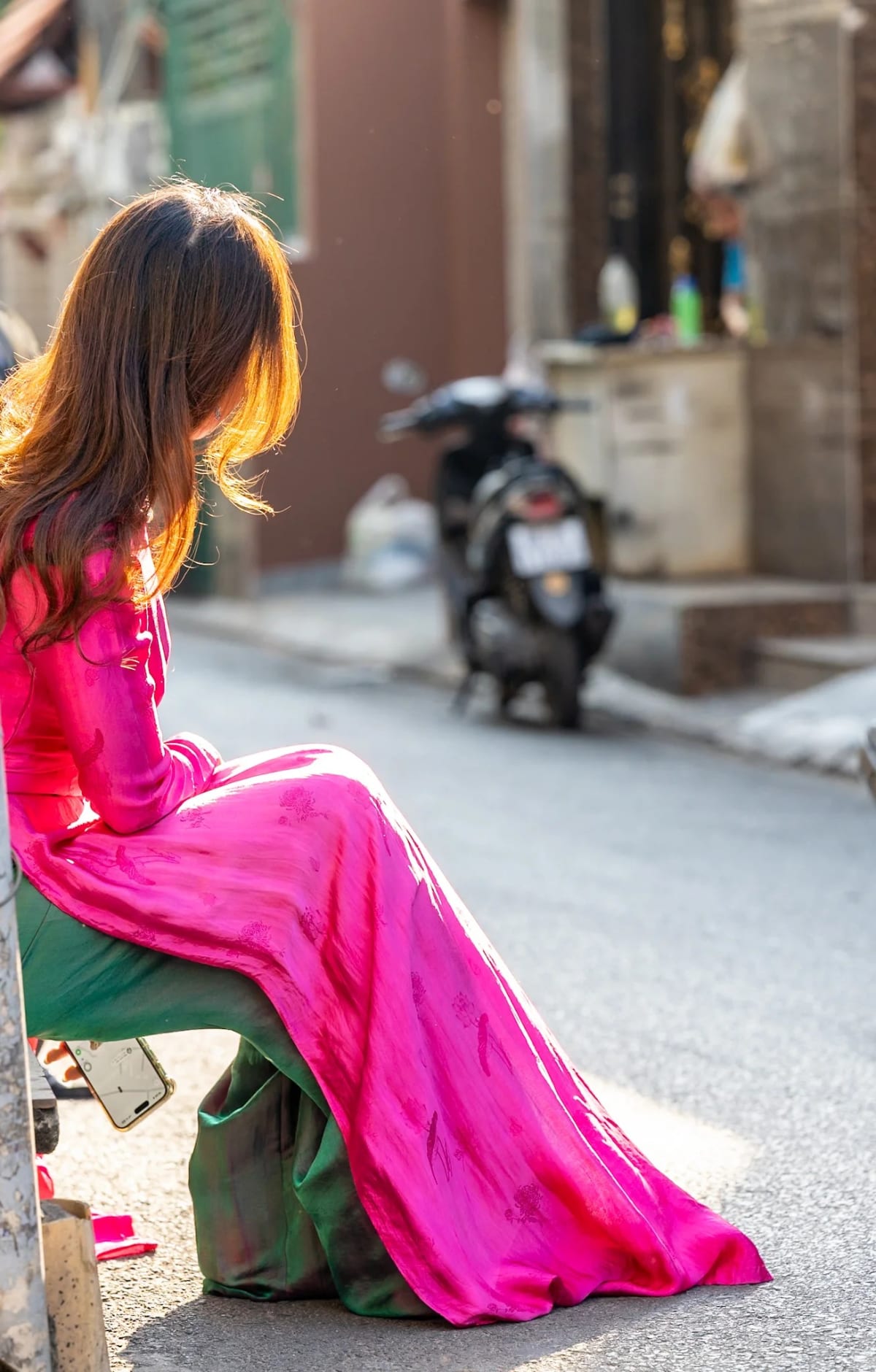 A woman in a pink áo dài sits at the roadside, her long hair lit from behind by the late afternoon sun — Jim Kerns