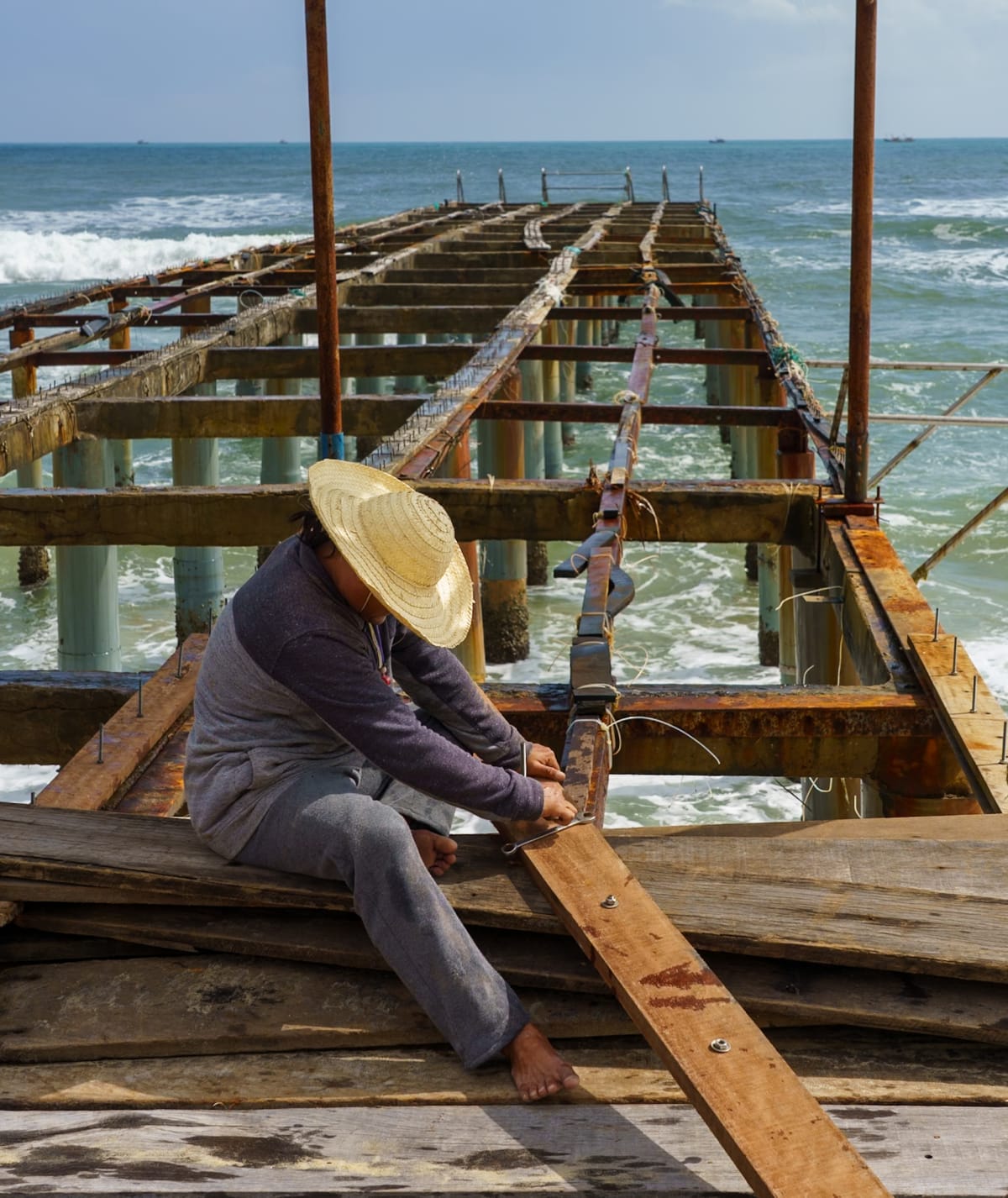 A man in a straw hat sits astride the timbers of a rusting pier, working a bolt as the sea breaks beneath him — Jim Kerns